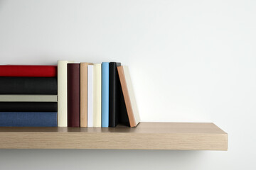 Many hardcover books on wooden shelf near white wall. Space for text