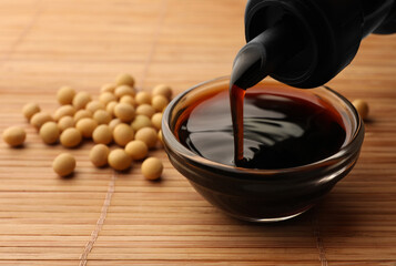 Pouring soy sauce from bottle into bowl and soybeans on bamboo mat, closeup