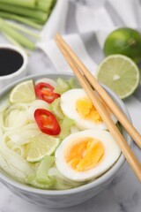 Bowl of delicious rice noodle soup with celery and egg on white marble table, closeup