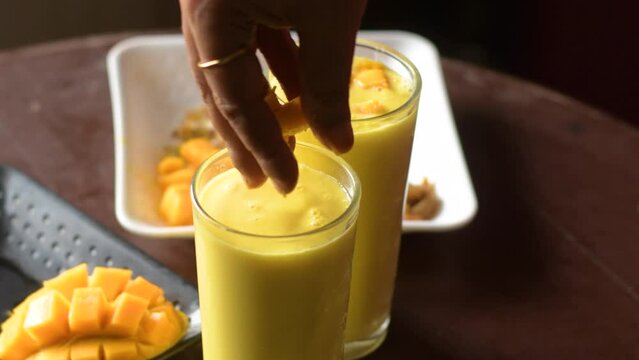 A lady putting mango slices and chopped nuts on top of mango lassi or mango yogurt smoothie a tasty popular drink in India.