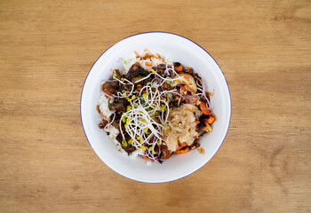 Vegan cuisine. Gluten free. Top view of a bowl with grilled tofu, soy sprouts, pumpkin seeds, season vegetables, spicy mango, sushi rice, cucumber and kimchi, in the restaurant wooden table.