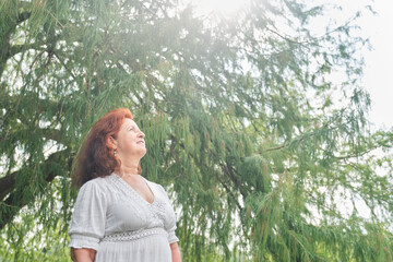 Portrait of a latin senior red-haired woman smiling looking up daydreaming in nature. Bright composition with copy space. Concept: peace of mind and happiness enjoying leisure time during retirement.