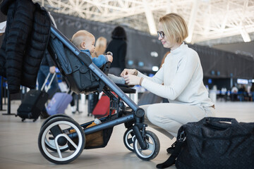 Obraz na płótnie Canvas Motherat interacting with her infant baby boy child in stroller while travelling at airport terminal station. Travel with child concept