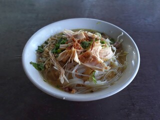 Isolated on brown wooden table a bowl of chicken soto or chicken soup.  Chicken soup Soto is a typical Indonesian food topping with shredded chicken. Selective focus