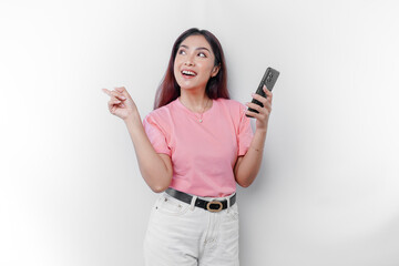 A portrait of a happy Asian woman wearing pink t-shirt, holding her phone, and pointing copy space on beside her, isolated by white background