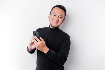 Excited Asian man wearing black shirt smiling while holding his phone, isolated by white background