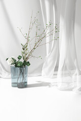 White rose bouquet and green tree twig in blue glass vase on counter table, soft blowing sheer fabric curtain in sunlight for luxury cosmetic, skincare, beauty, fashion product display background 3D