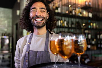 Happy smiling young waiter wearing apron standing in bar holding tray of beers looking at camera.
