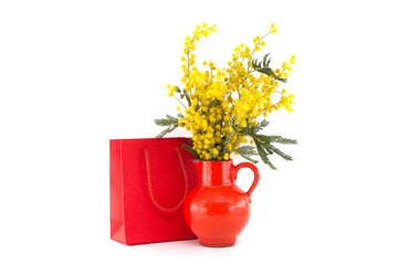 Bouquet of yellow mimosa flowers in red vase near red gift bag