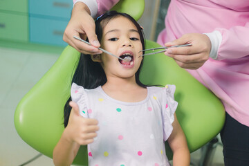 Little asian girl sitting at dental chair with open mouth during oral check up while doctor....