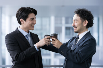 A boss (manager) and a subordinate businessman in suits toasting each other with coffee during a break.