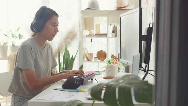 Professional young Asia lady IT development programmer listen music enjoy work type on keyboard coding programming fixing database on computer screen and laptop on table in workroom at house office.