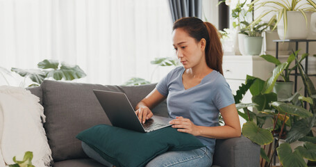 Obraz premium Young Asian woman wear casual calm sitting on wing chair use laptop computer thinking the idea for working full of plants in living room greenhouse. Coronavirus and stay home, Work from home concept.