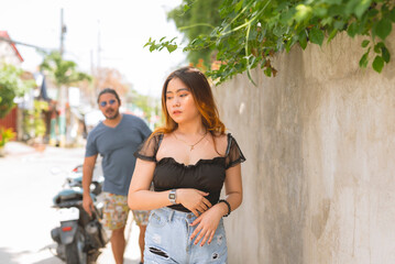 A pretty young lady is on the roadside looking sideways has her back turned against an older guy in sunglasses who seems to be checking her out. A motorbike, road and poles in the background.