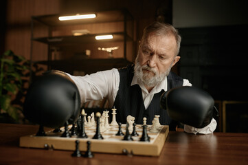 Senior man chess player moving chesspiece on board while wearing in boxer gloves