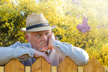 Ein älterer Mann mit Hut steht hinter einem Gartenzaun. Er stützt seinen Kopf auf eine Hand und...