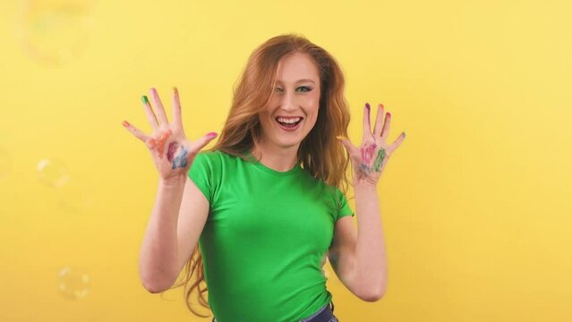 A girl dancing on an orange background shows her painted hands to the camera. A cheerful red-haired woman dances in front of the camera while soap bubbles fly around her.