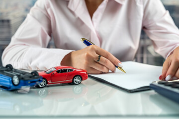 woman is writing  document about car insurance at table next to  car model and a calculator.