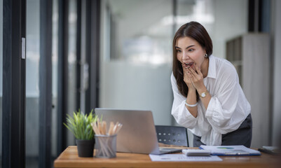 Excited Happy Asian young woman using phone and laptop sitting on a desk office in the day at office	