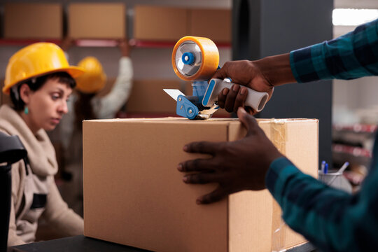 Delivery office manager packing parcel using hand held sticky tape dispenser. African american shipping department worker arms closing cardboard box with adhesive close up
