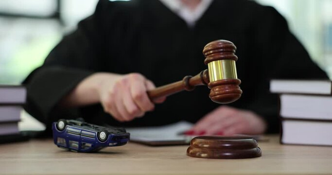 Female judge in mantle knocks wooden gavel sitting at table with overturned toy car . Accident and litigation concept in court slow motion