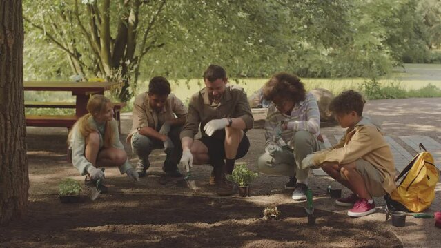Male scout leader using small shoulder blade while showing how to plant flowers in soil at public park to diverse group of school children