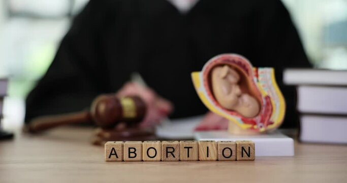 Word Abortion with wooden cubes on judge counter in office. Judge takes notes to prosecute next to fetal model. Concept legislation against abortion slow motion