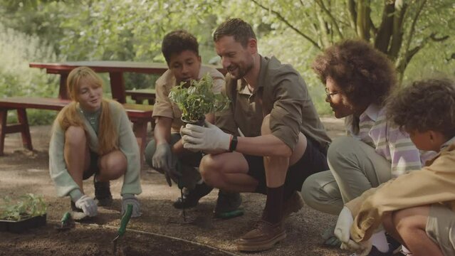 Male ranger holding potted plant in hands while speaking to multiracial group of volunteer children taking part in environmental planting project in public park