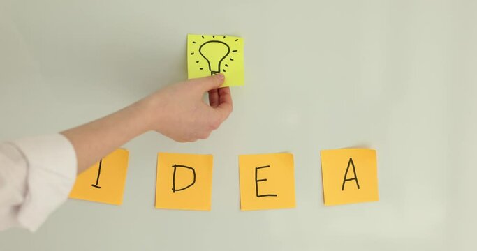 Woman sticks yellow paper with glowing light bulb image over inscription Idea on white wall. Concept of development and new flashing intentions slow motion