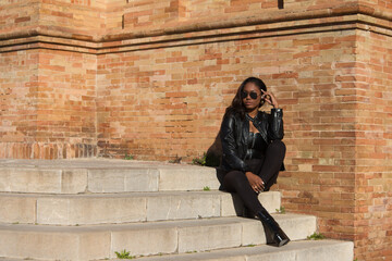 young and beautiful black latin woman wearing black clothes and sunglasses is sitting on the steps of the most important square in the city of seville, spain. The photo is taken from the front.