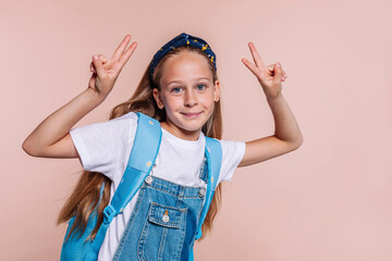 Blond school girl with blue eyes, wears a blue backpack and denim overalls, does peace victory sign...