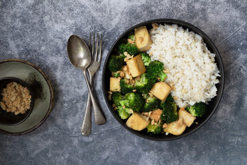 Vegan healthy food stir fried broccoli tofu peanuts has brown rice in black bowl on slate table.