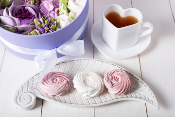 Pink and white marshmallows on a wooden saucer next to a cup of tea and a box of marshmallow flowers