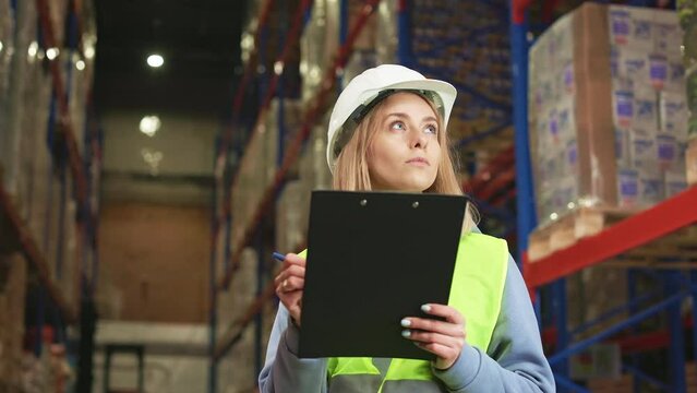Concentrated caucasian woman taking notes on clipboard while checking goods and supplies on shelves in warehouse. Female inspector in working uniform doing regular inventory for data updating.