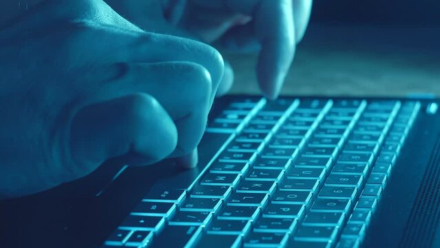 Man's Hands Typing On A Black Laptop With Illuminated Keyboard Lying On A Desktop On Dark. Closeup. Shallow Depth Of Field