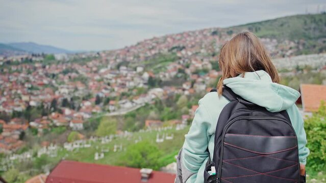 A young adult Caucasian woman taking pictures of the city of Sarajevo from a viewpoint with a smartphone while traveling to the Balkans.