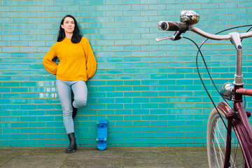 Thoughtful young woman in casual clothes leaning near skateboard on turquoise wall