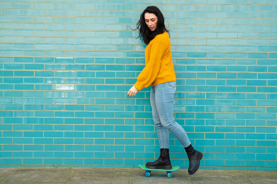 Young woman in casual clothes skateboarding in front of turquoise wall