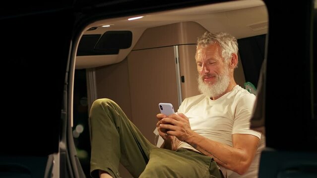 Older man sitting inside rv camper van with tent on it on vacation using mobile phone. Senior mature traveler holding smartphone enjoying free internet or checking rentals in camping at night.