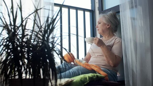 A pensive elderly woman sits alone at the window, drinks morning coffee, enjoys the warm weather.
