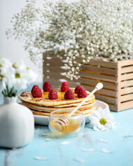 Pancakes with honey and berries for breakfast. Honey in bowl with honey dipper on table with white flowers in wooden box. Wooden blue background. Side view. 