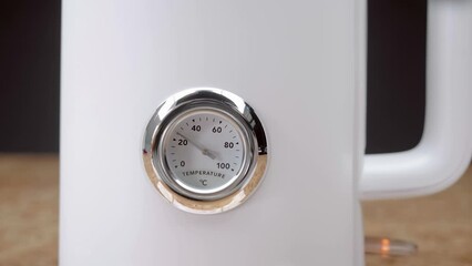 A white electric kettle with a round thermometer on which the arrow moves to a temperature of 100 degrees Celsius, boils and steam escapes from the spout of the device on a black background