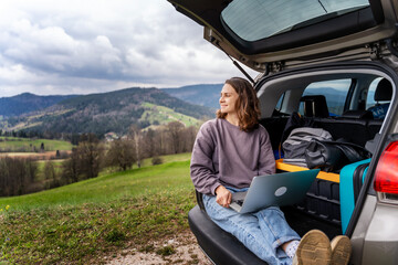 Young pretty cheerful woman traveler sitting in car trunk using laptop while traveling in mountains - 602530753