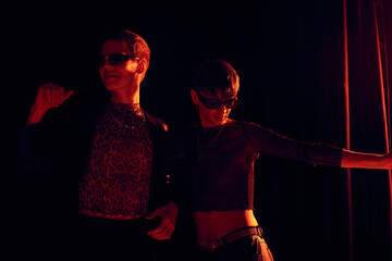 Fashionable nonbinary friends in party outfits and sunglasses dancing together while celebrating lgbt pride month on black background with red lighting