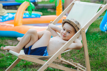 A little boy is resting in the garden near the house on a wooden deck chair in summer