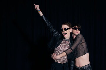Stylish and young gay man in shiny top hugging excited queer friend in sunglasses and trendy outfit during lgbt pride month and party isolated on black
