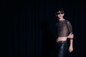 Trendy young gay man in sunglasses, sparking top and leather pants posing while celebrating lgbt community and pride month isolated on black