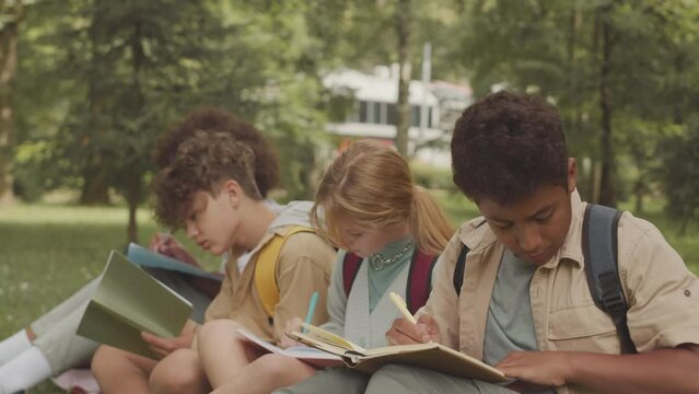 Group of multicultural elementary school children writing in their copybooks while sitting on grass in public park having outdoor class