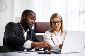 Fototapeta premium Two people analyze documents on a laptop while sitting at a table in the office. Business partners at work in the office are discussing projects. Multi-racial collaboration.