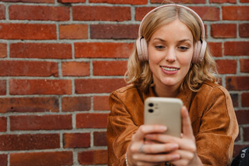 Smiling woman listening music with headphones and mobile phone while sitting outdoors near brick wall